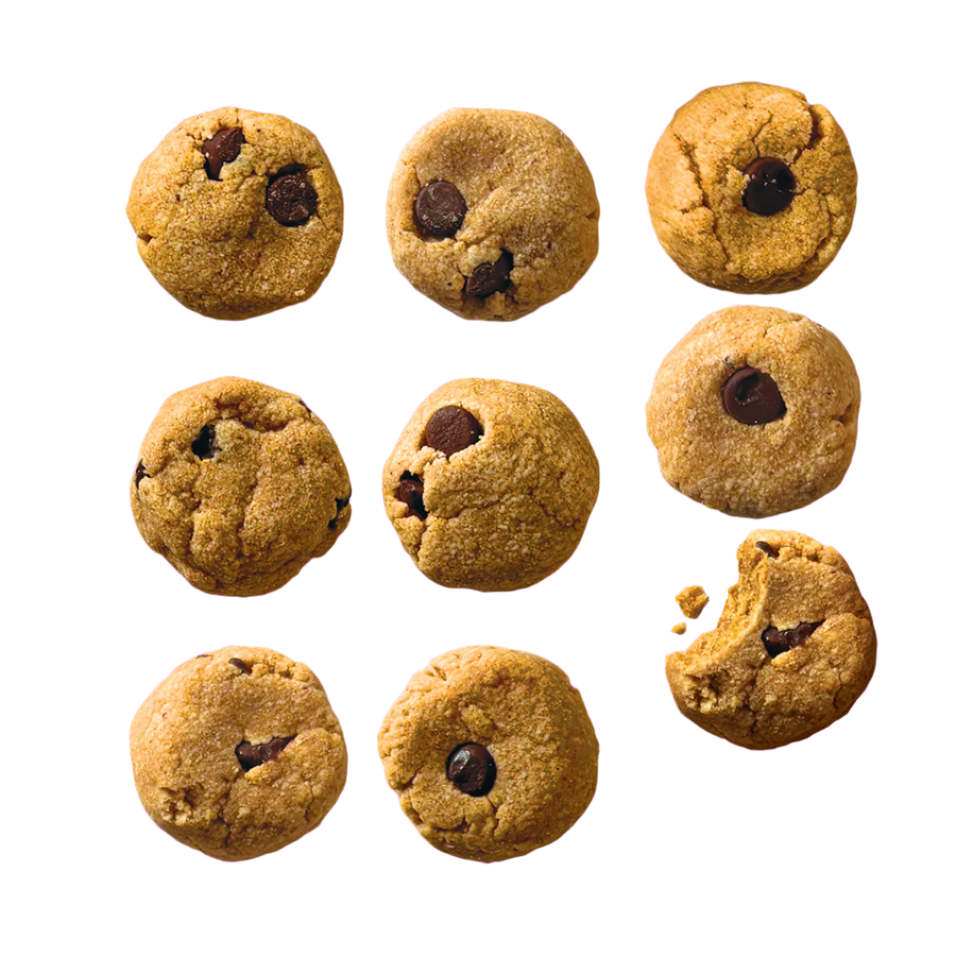 Nine soft-baked, golden-brown cookies, each studded with upcycled date "chocolate chips", are artfully scattered across a bright surface. One cookie in the bottom right corner has a bite taken out of it, revealing its moist, light interior. Each cookie is a perfect bite size morsel of these tasty treats!